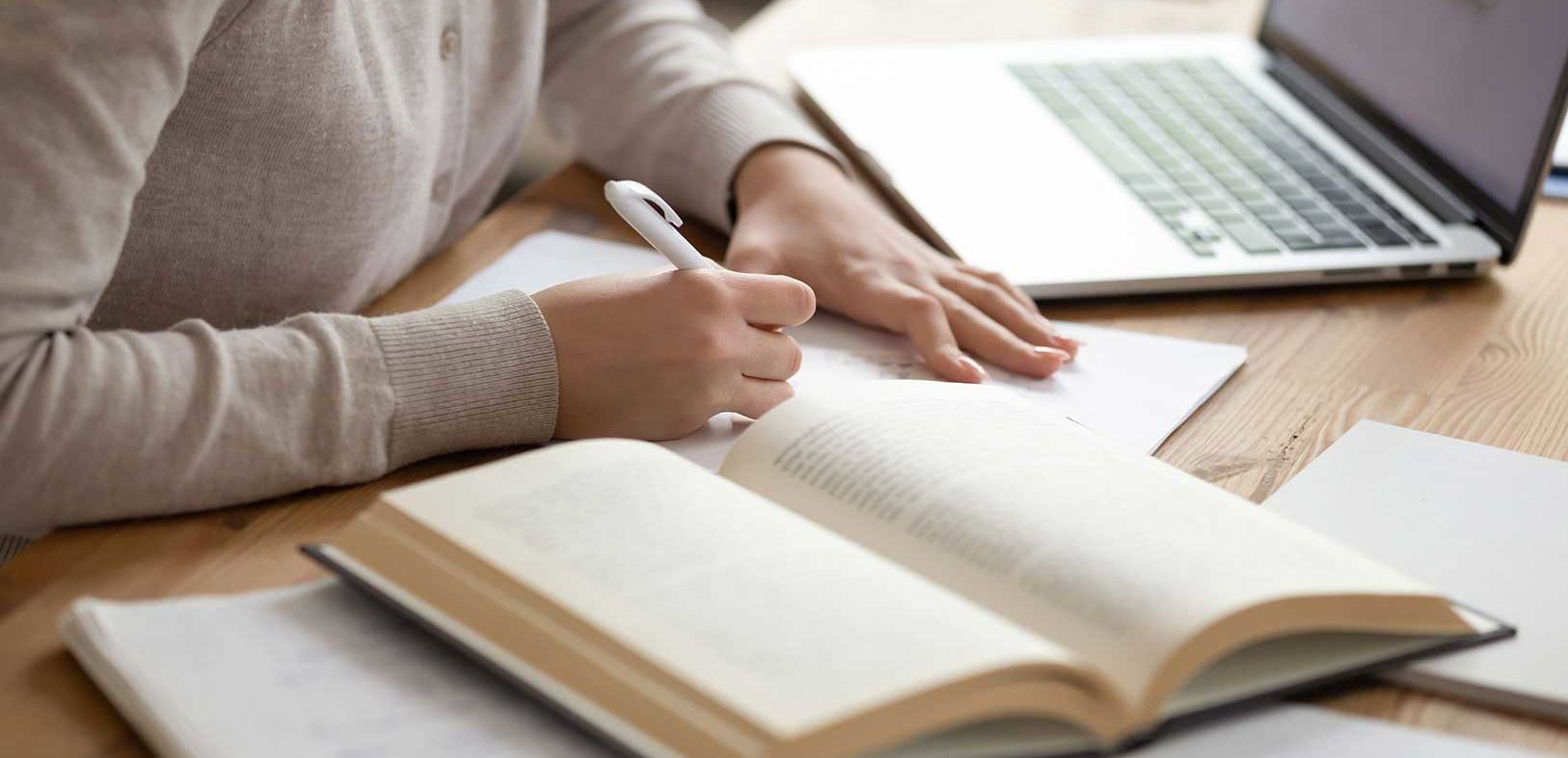 A person writing on paper surrounded by a laptop and open books