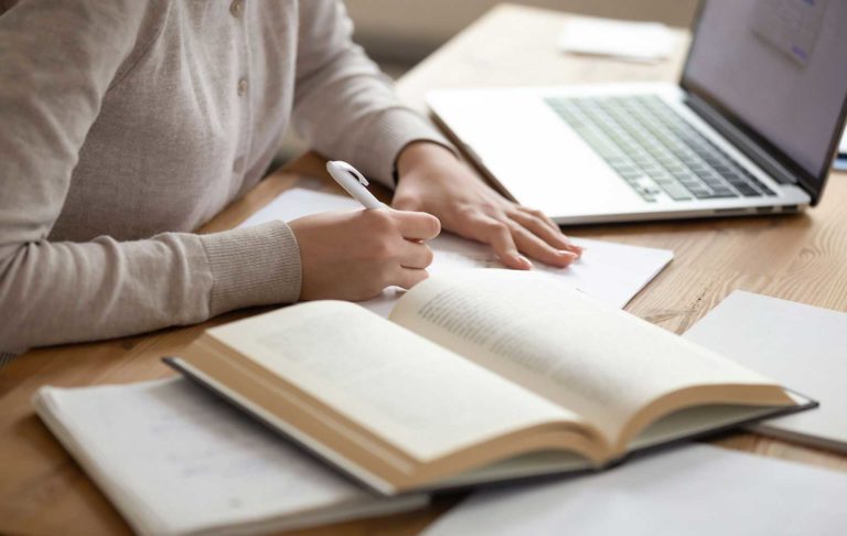 A person writing on paper surrounded by a laptop and open books