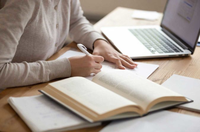 A person writing on paper surrounded by a laptop and open books
